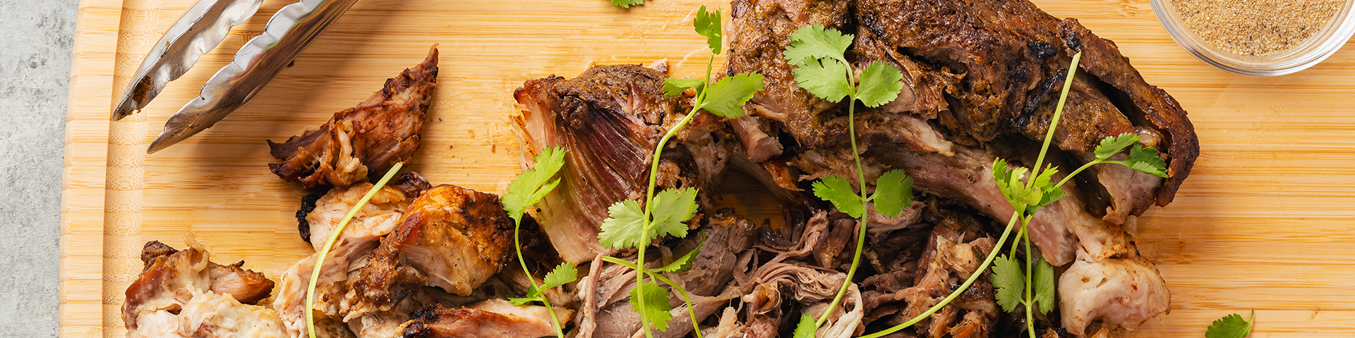top view of a cuban bbq pork roast shredded on a wooden cutting board. On top of the pork roast is fresh cilantro. Tot he side of the pork roast is an orange cut in half, a pinch bowl with Rodelle's cuban seasoning, and a silver pair of tongs. The background is a neutral mid gray