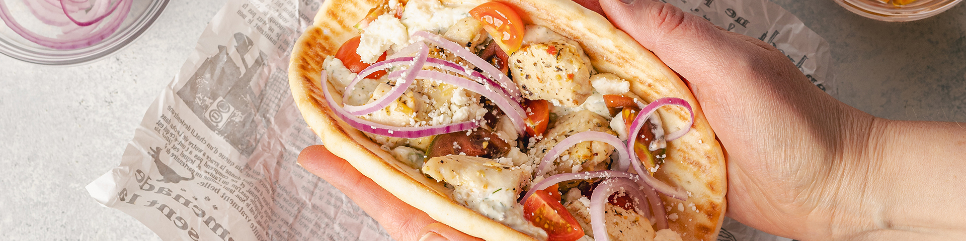 Top view of a plate with greek chicken gyros assembled. On a large pita bread are tomatoes, greek seasoned chicken, onion, feta, and a homemade tzatziki sauce. Surrounding the gyro which is on some parchment paper are all the ingredients to assemble the gryo. The background is white.