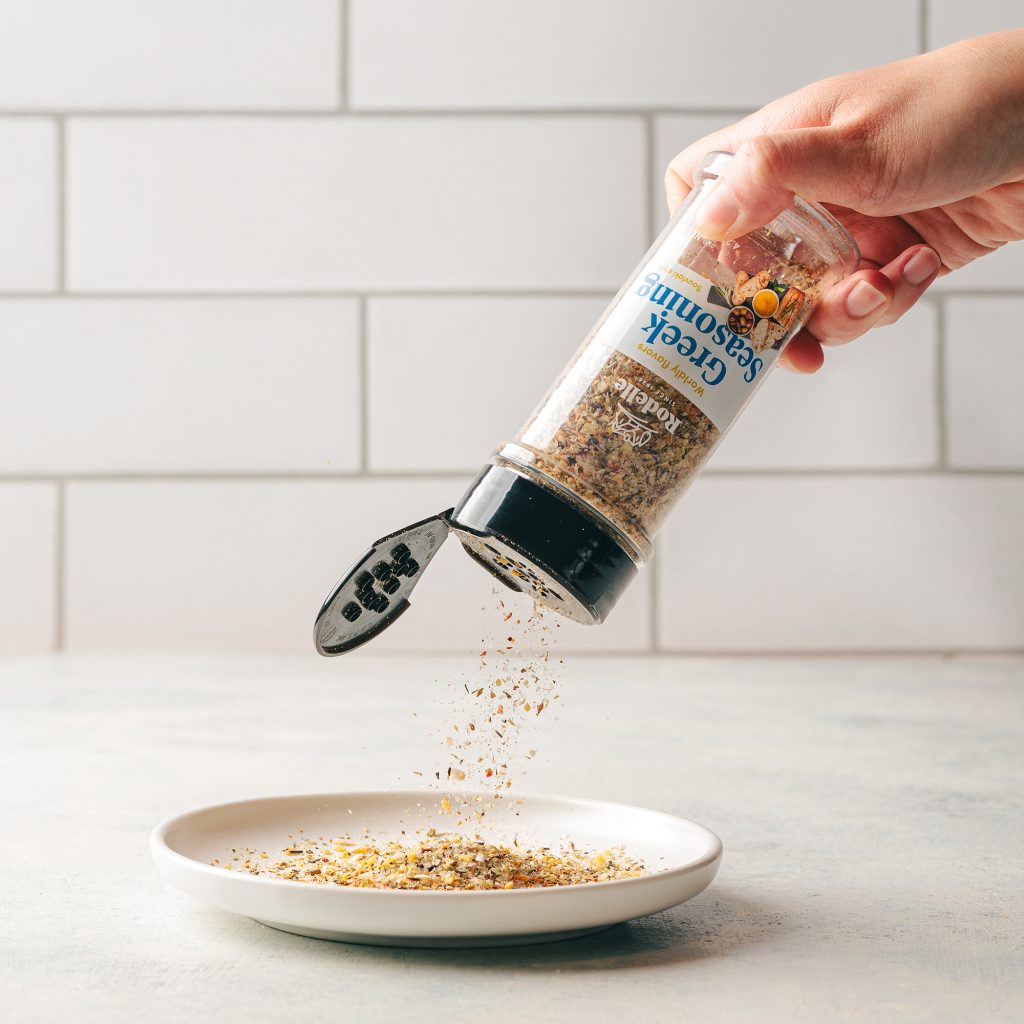 lifestyle shot of Rodelle's 4.25oz Greek Seasoning being poured onto a plate. The bacgkround is white tile and the table is a light marble gray pattern.