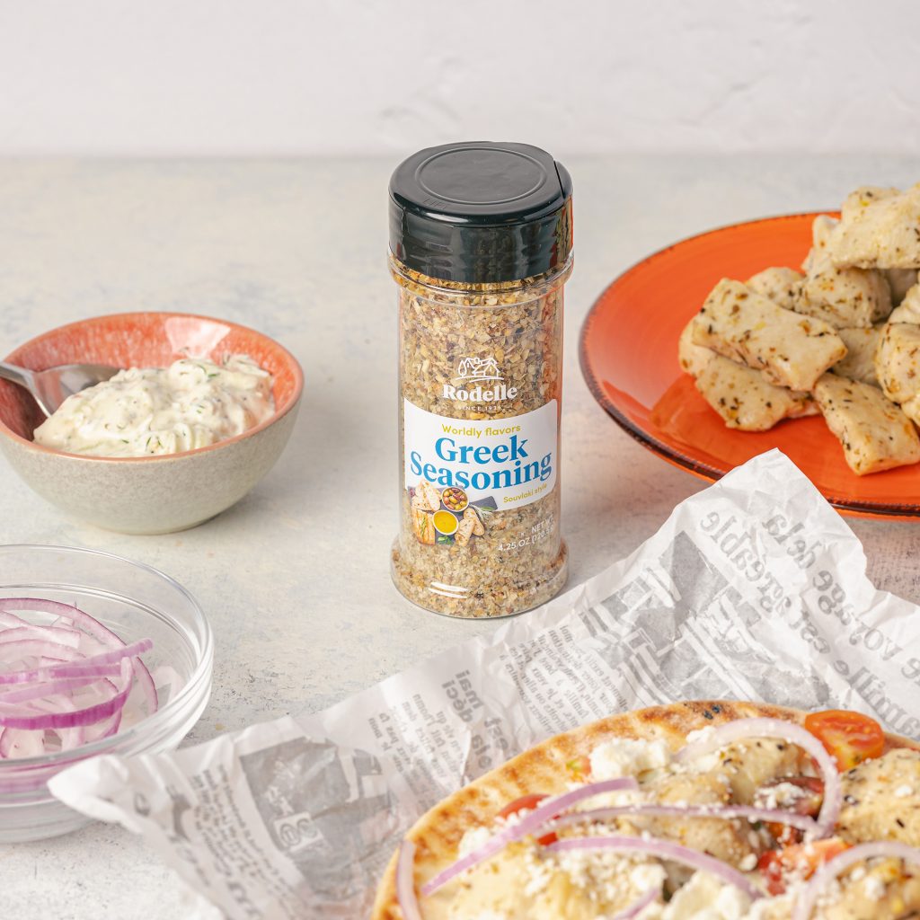 lifestyle image of Greek Chicken Gyros With Tzatziki Sauce laying on a white parchment paper. Rodelle's greek seasoning lays at the center and surrounding it was a vibran orange plate with greek seasoning chicken. In a orange and earth toned bowl is homemade tatziki sauce to the upper left and bottom left is a clear sauce bowl with slices of red onion