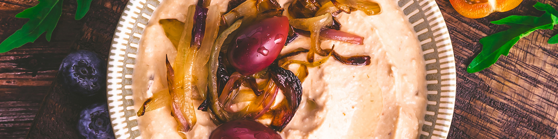 Whipped feta in a white bowl with grilled onions and tomatoes used for garnish on top. Surrounding the whipped feta is crusty bread, basil, tomatoes, grilled onions, and much more. Everything lays on a wooden table