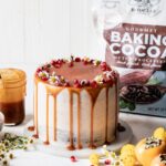 Brownie layer cake with caramel drip and seasonal fall fruits on a neutral background with Rodelle cocoa in back