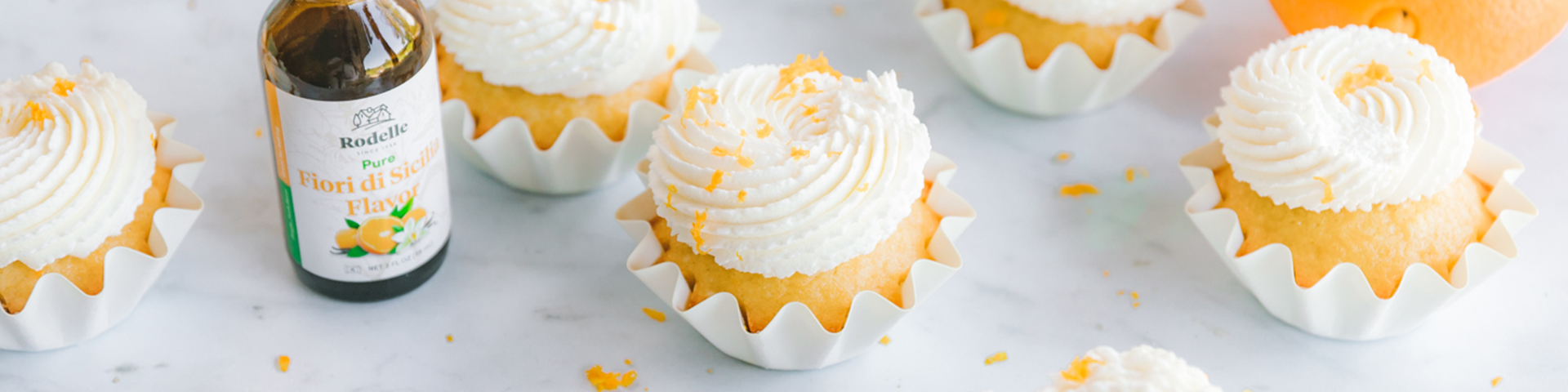 fiori di sicilia cupcakes with piped whipped cream frosting, laying in white cupcake tins, garnished with orange peel. Rodelle pure fiori di sicilia flavor in scene. Whole oranges in background