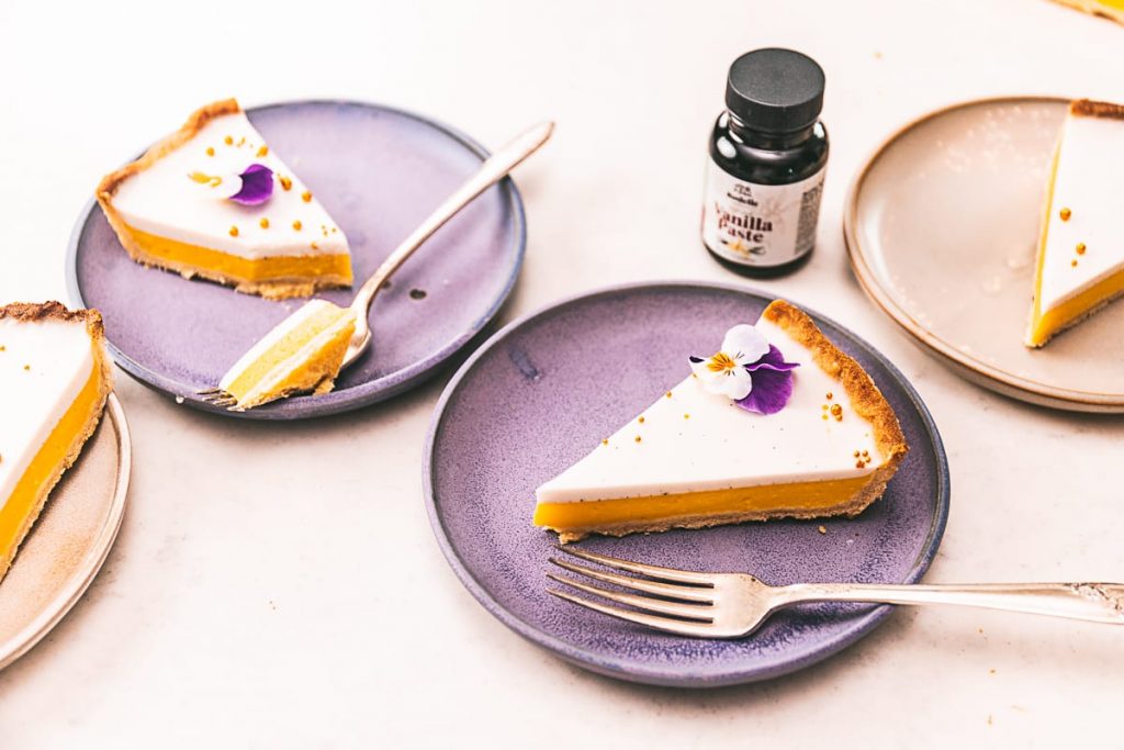 Mango tart with vanilla coconut custard sliced on neutral background with edible flours on purple plates