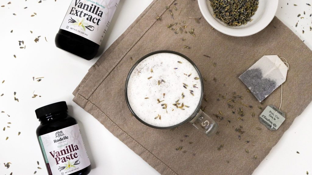 Earl grey tea with vanilla milk foam in clear glass mug on neutral background with dried lavendar and Rodelle products