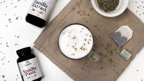 Earl grey tea with vanilla milk foam in clear glass mug on neutral background with dried lavendar and Rodelle products