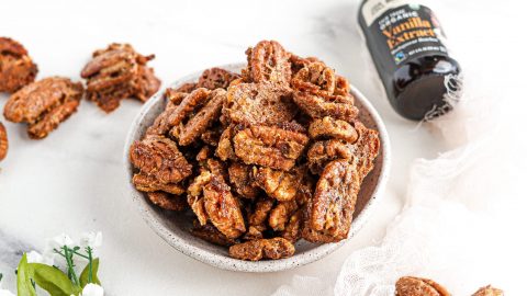 Caramelized pecans in a white bowl with vanilla extract in background