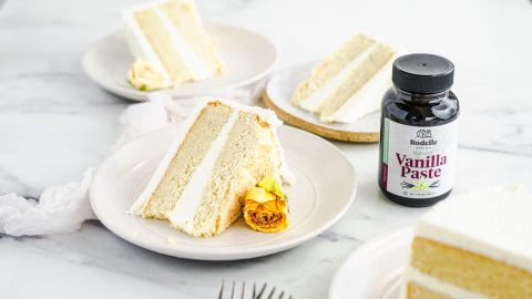 Three pieces of vanilla cake with vanilla frosting on white plates and marble counter showing Rodelle Vanilla Paste