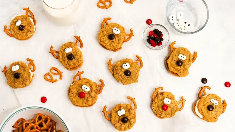 Close up top shot of a large group of oatmeal cream pie reindeers with red and brown m&m noses and reindeer antlers made with pretzels. Around the scene are pieces of m&ms, a bowl of m&ms, broken pretzels, pretzels in a bowl, handmade candy eyes, and a glass of milk