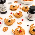 angle shot of a group of oatmeal cream pie reindeer cookies with pretzels for antlers, handmade candy eyes, and red and brown m&ms for button noes. In the scene are pretzels and m&ms scattered around the scene and Rodelle vanilla paste and rodelle pure vanilla extract.