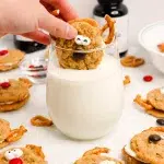 Hand holding an oatmeal cream pie reindeer cookie and dunking it in a clear glass of milk. Surrounding the scene are other cookies