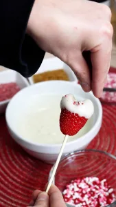 hand sprinkling pink, red, and white sprinkles on a chocolate covered heart in shape of a heart on a wooden skewer.