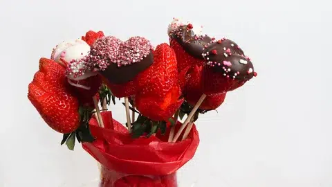 A mix of white and milk chocolate covered strawberry hearts and strawberry roses in a glass vase with red paper.