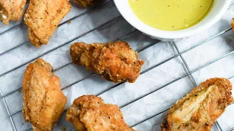 Close up of cuban BBQ wins with mojo dipping sauce in a white bowl. Food laying on top of a cooling rack with parchment paper below.