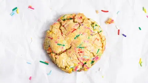 Centralized shot of single-serve funfetti cookie on crinkled white parchment paper. Sprinkles randomly dispersed around cookie.