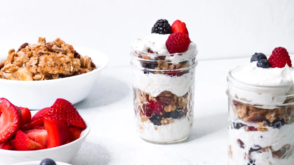 vanilla berry granola parfait in 6oz mason jars. white bowls of fruit, granola on side.
