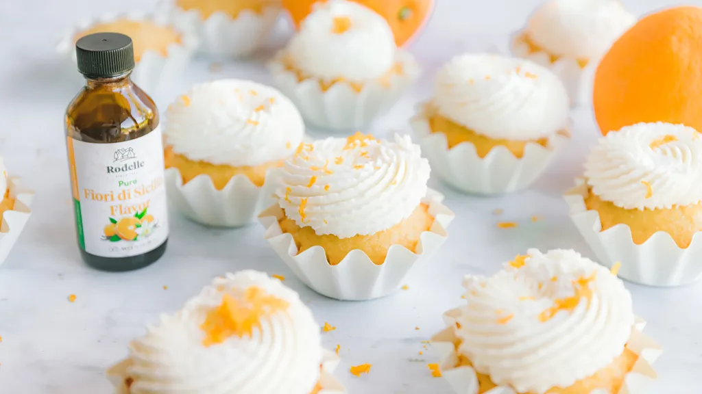 fiori di sicilia cupcakes with piped whipped cream frosting, laying in white cupcake tins, garnished with orange peel. Rodelle pure fiori di sicilia flavor in scene. Whole oranges in background