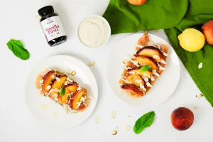 top view of two pieces of peach and prosciutto toast with vanilla whipped ricotta spread sitting on top or white plates. In the upper left corner is Rodelle's 4oz Natural Vanilla Paste and the spread in a small white glass bowl. To the upper right is a green towel with peaches and lemons laying on top. Bottom right has sliced almonds and 1 large mint leaf.