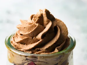 close up of piped cocoa whipped cream in a clear glass jar with a light grey textured background