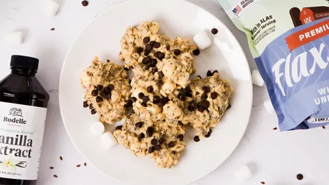 Top view of no bake rocky mountain cookies with mini chocolate chips sprinkles on top. Scattered around are marshmallows and chocolate chips. Rodelle Pure Madagascar Bourbon Vanilla Extract and Knwble Grwn Flax Seeds lay on the side.