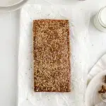 Top view of a whole halva (5in x 9in) piece baked laying on parchment paper. Surrounding the scene are white plates with rectangular wrapped pieces of halva, and a small sauce white glass dish containing sesame seeds.