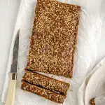 Top view of a large rectangular halva piece being cut into slices so they are bar shaped. Surrounding the halve is a cutting knife, parchment paper, and white plate with rectangular shaped halva wrapped in parchment paper.