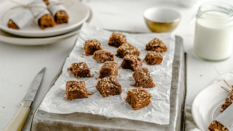 Homemade halva pieces cut into cubes laying on a sheet pan with parchment paper. Surrounding the halva is a cup of milk and white plates with wrapped up halve pieces