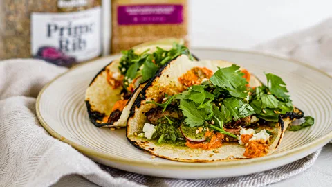 Close of two prime tacos containing steak pieces, homemade salsa verde, toasted pepita salsa rojo, and topped with diced onion and cilantro. Tacos lay on a light beige plate with a beige napkin surrounding them. In the background is also Rodelle Prime Rib Seasoning and Rodelle Toasted Sesame Seeds.