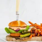 Side profile of a turkey burger containg tomato, red onion, a light orange sauce, The burger has been stabbed with a knife and sits on top of parchment paper, and a wooden board. Next to the burger are sweet potato fries.