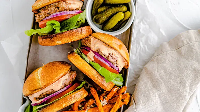 Top view of turkey burgers on their side in a small sheet pan with a bowl of small pickles and sweet potato fries.