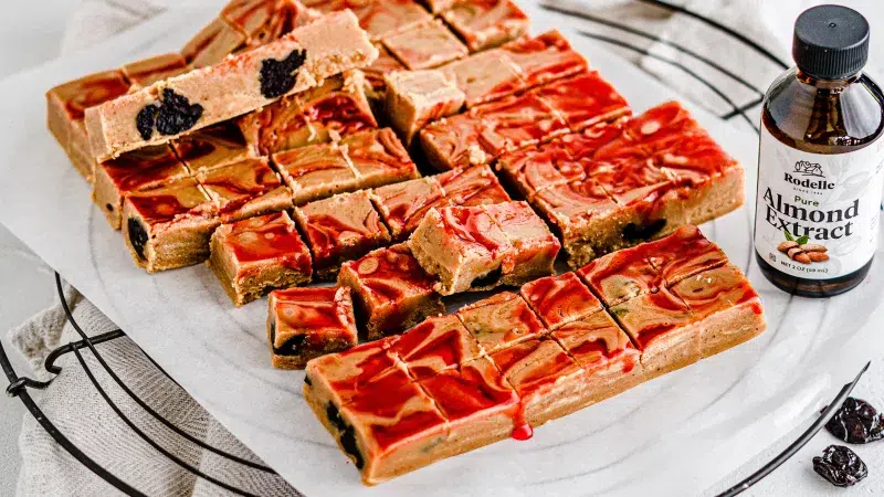 Top angle shot of cherry amaretto sidecar fudge sliced up. They lay on white parchment paper and a black circular cooling rack. Next to the fudge is Rodelle's Almond Extract.