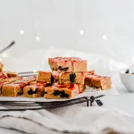 Side view of cherry amaretto sidecar fudge. Some of the fudge is stacked on top. It lays on parchment paper and a black circular cooling rack. Surrounding the fudge are small bowls and white with thin gray striped towel.