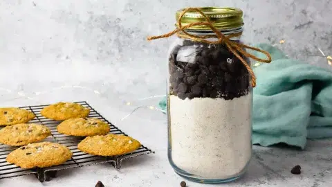 A side profile of a 16oz jar containing a diy cookie mix with a top layer of chocolate ship cookies. The mason jar has a gold lid on top and a bow made out of twin wrapping the top portion for decorations. behind the jar is a light blue towel and to the left is a cooling rack with chocolate chip cookies. Chocolate chips surround the the photo and are scattered.