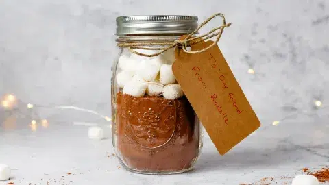 Side profile of a homemade diy hot cocoa mix in a jar with marshmallows. Twine holds a kraft paper take that reads "To: Our Friend" and "From: Rodelle."
