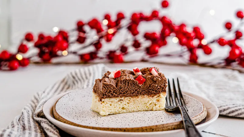Side profile view of a peppermint sugar cookie bar on a textured plate with a fork. Surrounding the plate is a light tan towel and in the background are branches with red berries.