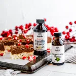 Side profile photo of peppermint sugar cookie bars laying on parchment paper that's on a cookie tray. Around the sugar cookies is Rodelle's Peppermint Extract and Alcohol Free Vanilla Flavor. In the background of red berries on branches.
