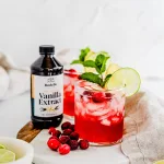 Side view of the vanilla cranberry apple mule. There are two glasses served with both drinks and containers. Whole cranberries lay nxt to the drinks and Rodelle's 8oz Gourmet pure