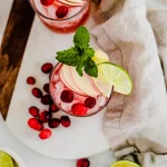 top view of vanilla cranberry apple mule. Two glasses are in the scene containing two slices of lime and mint. On the ground is a tan kitchen towel. The glasses lay on a wooden