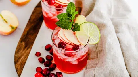 Top angle view of Vanilla Cranberry Apple Mule laying on a wooden surface with a cool tan towel. In the scene are crangerries next to them. In the vanilla cran apple mule are two slices of lime and mint.