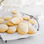 Close up of round vanilla peppermint shortbread dipped in white chocolate that are laying on white parchment paper and a black round wired cooling rack. The background is white with texture and there are fairy lights blurred in the background.