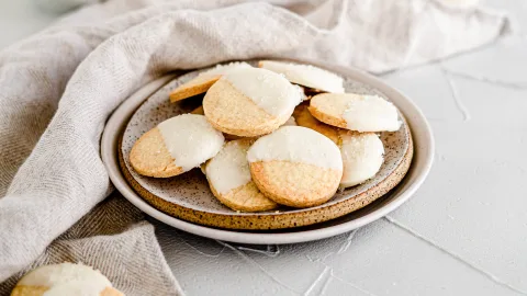 Landscape photo of a bundle of Vanilla Peppermint Shortbread dipped in White Chocolate round cookies laying on a white with brown speckled plate. Surrounding the cookies is a gray kitchen cloth.