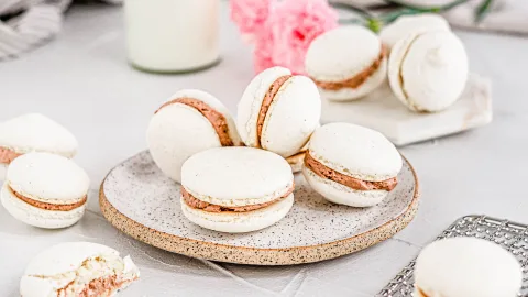 close up of vanilla macarons with chocolate hazelnut filling on a textured clay plate. The background is white and contains a pink flower.