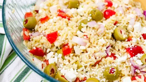 Close up of greek pasta salad in a large clear bowl containing orzo pasta, red bell peppers, green olives, and onions.