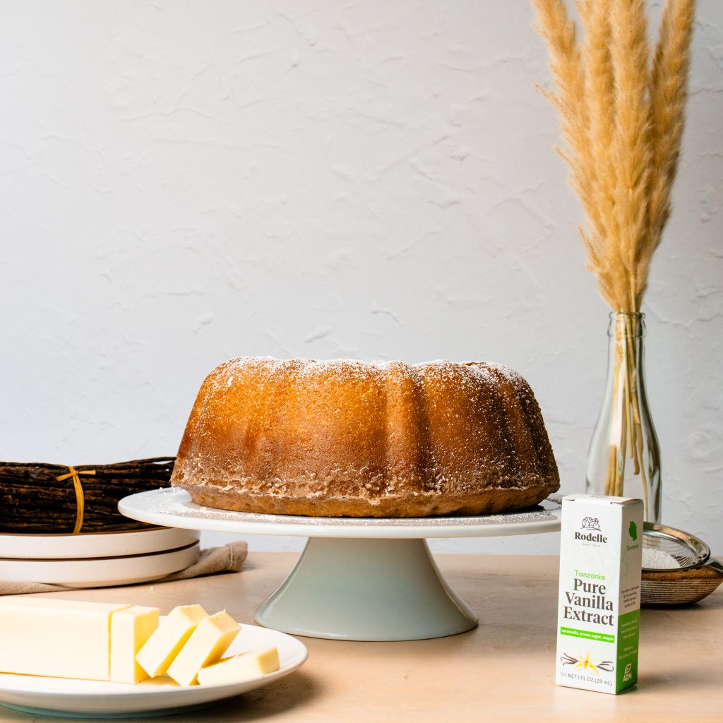 recipe image of a kentucky butter bundt cake on a white cake stand in the center. surrounding the image is a plate with butter - sliced, and in the bottom right is Rodelle 1oz tanzania pure vanilla extract in a box. the background is a neutral gray/white.