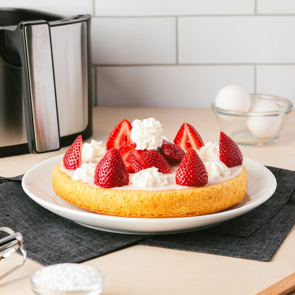 recipe photo of a vanilla airfryer cake with dollops of whipped cream and strawaberries on top. The cake is on a white plate and underneath the plate is a dark black towel. n the background of the cake is white tile and an airfryer.