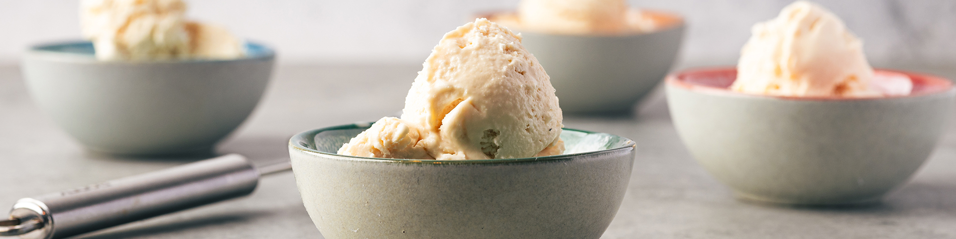 Close up of scoops of no churn vanilla ice cream in small ice cream bowls with a blue-ish green exterior but color interior including, green, pink, orange, red. There is one ice cream bowl up front and three in the background. A pile of vanilla beans are at the bottom right and a silver ice cream scoop in the background. The back drop and table are gray.