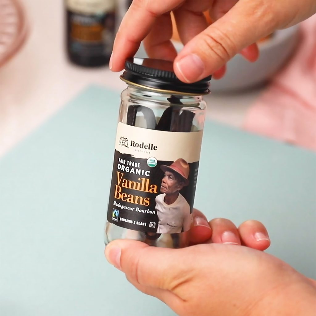 lifestyle image of two hands holding a Rodelle 3ct organic fairtrade vanilla beans jar in front of the camera. In the background is a light blue cutting mat.
