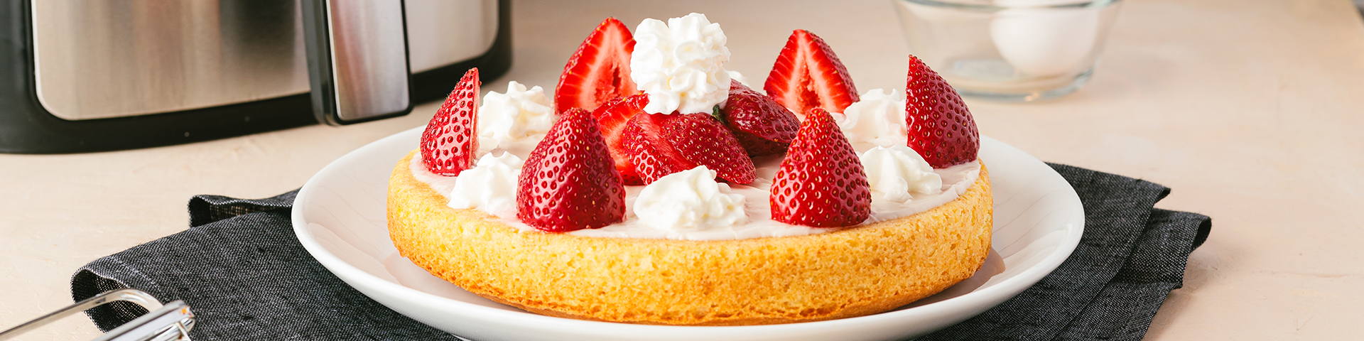 Top photo of an air fryer vanilla cake topped with a vanilla glaze, whipped cream, and fresh strawberries cut in half.