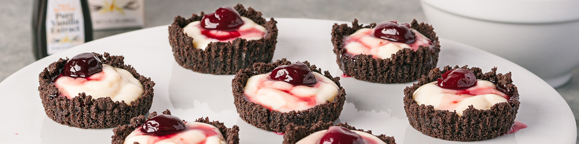 close up of seven cherry cordial tartlets on a white cake stand. In the background is Rodelle's bottle in a box, papua new guinea vanilla extract and a white bowl containing cherry syrup. On the gray textured table are crumbled pieces of oreo.