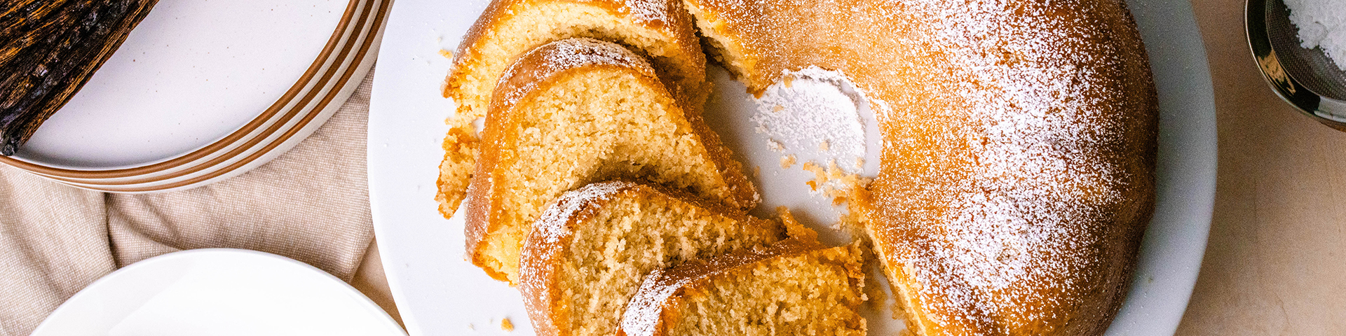 top view of a kentucky butter bundt cake on a white cake stand. The cake is dusted with powdered sugar and a portion of it is sliced and laying sideways. Surrounding the bake is a small plate with cut up butter, a bundle of vanilla beans, plates, and a sifter with powdered sugar. The table is a light pink and tan color